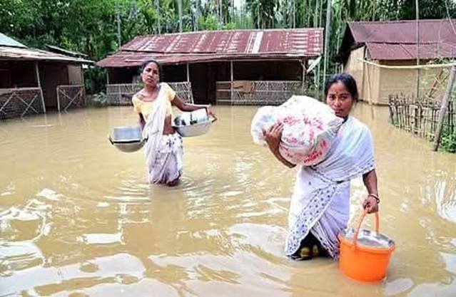 Two women wade through deep floodwaters carrying their belongings — displaced by the Assam floods