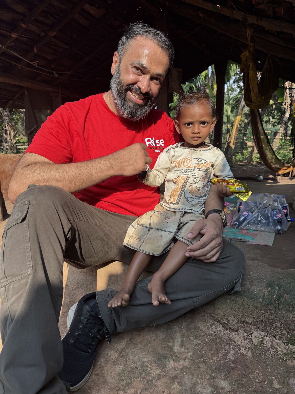 A RAHI team member holding a young child during a village visit under a traditional thatched shelter
